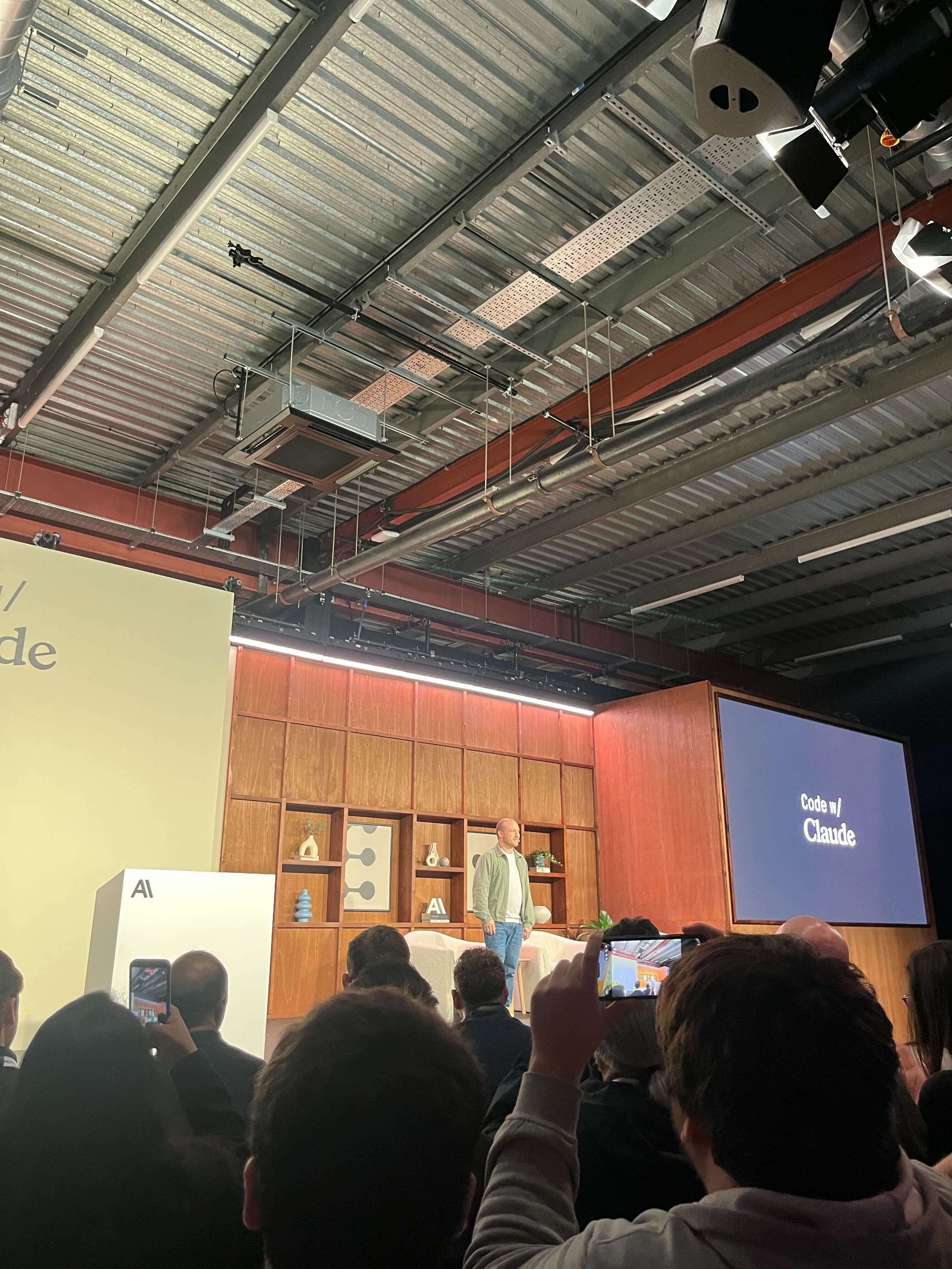 Speaker on stage at Code with Claude London, with a wooden Scandinavian-style set behind and the audience holding up phones in the foreground